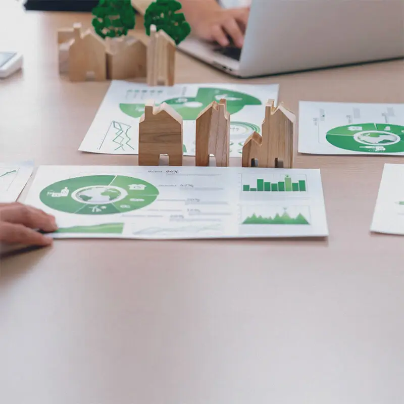Documentos con gráficos verdes sobre sostenibilidad y energías renovables en una mesa de trabajo. Maquetas de casas de madera refuerzan el concepto de planificación urbana sostenible y desarrollo ecológico.