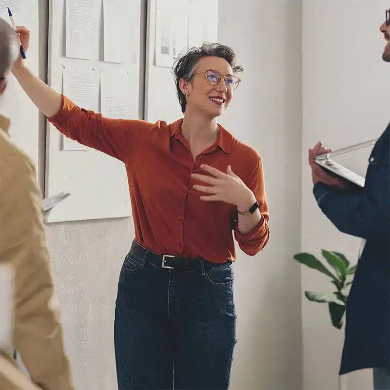 Mujer presentando una idea en una reunión de equipo. Con una actitud positiva, explica un concepto en un tablero de notas mientras sus colegas escuchan atentamente. Escenario de colaboración y liderazgo en un entorno empresarial moderno.