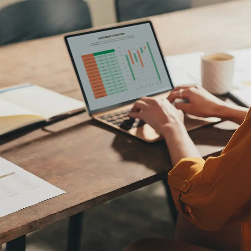 Mujer trabajando en un portátil con gráficos de inversión en pantalla. Un entorno de trabajo productivo con café y documentos en la mesa, reflejando análisis financiero y toma de decisiones.