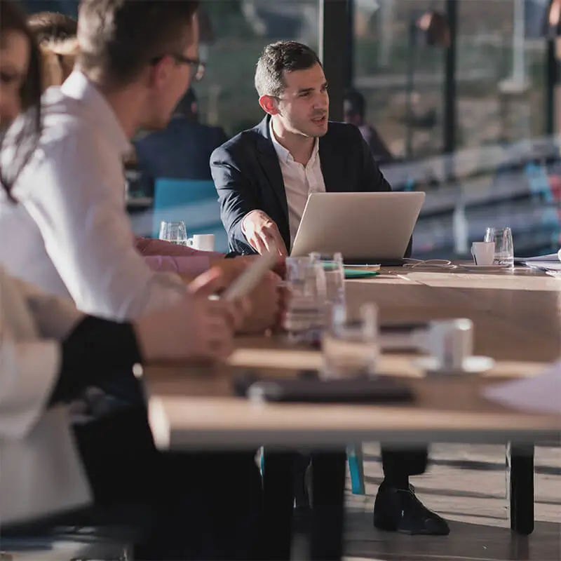 Ejecutivo liderando una reunión en una oficina moderna, con un portátil sobre la mesa y sus compañeros participando en la discusión. Representa liderazgo, trabajo en equipo y planificación estratégica.