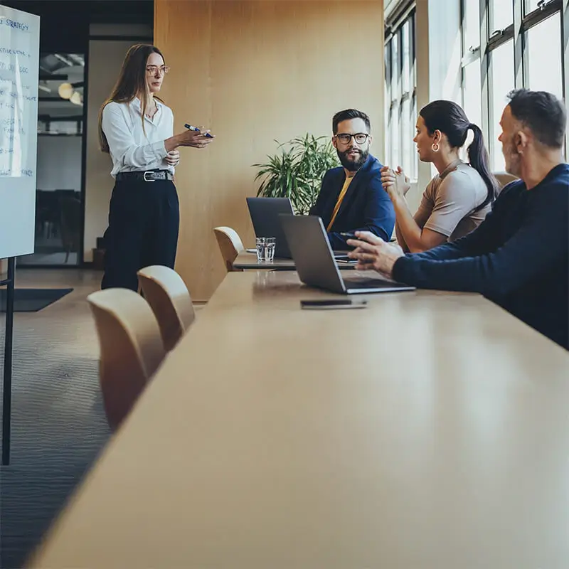 Reunión empresarial en una sala de conferencias moderna. Una mujer de pie presenta un informe mientras sus colegas la escuchan con interés. Conceptos clave: estrategia corporativa, liderazgo y toma de decisiones