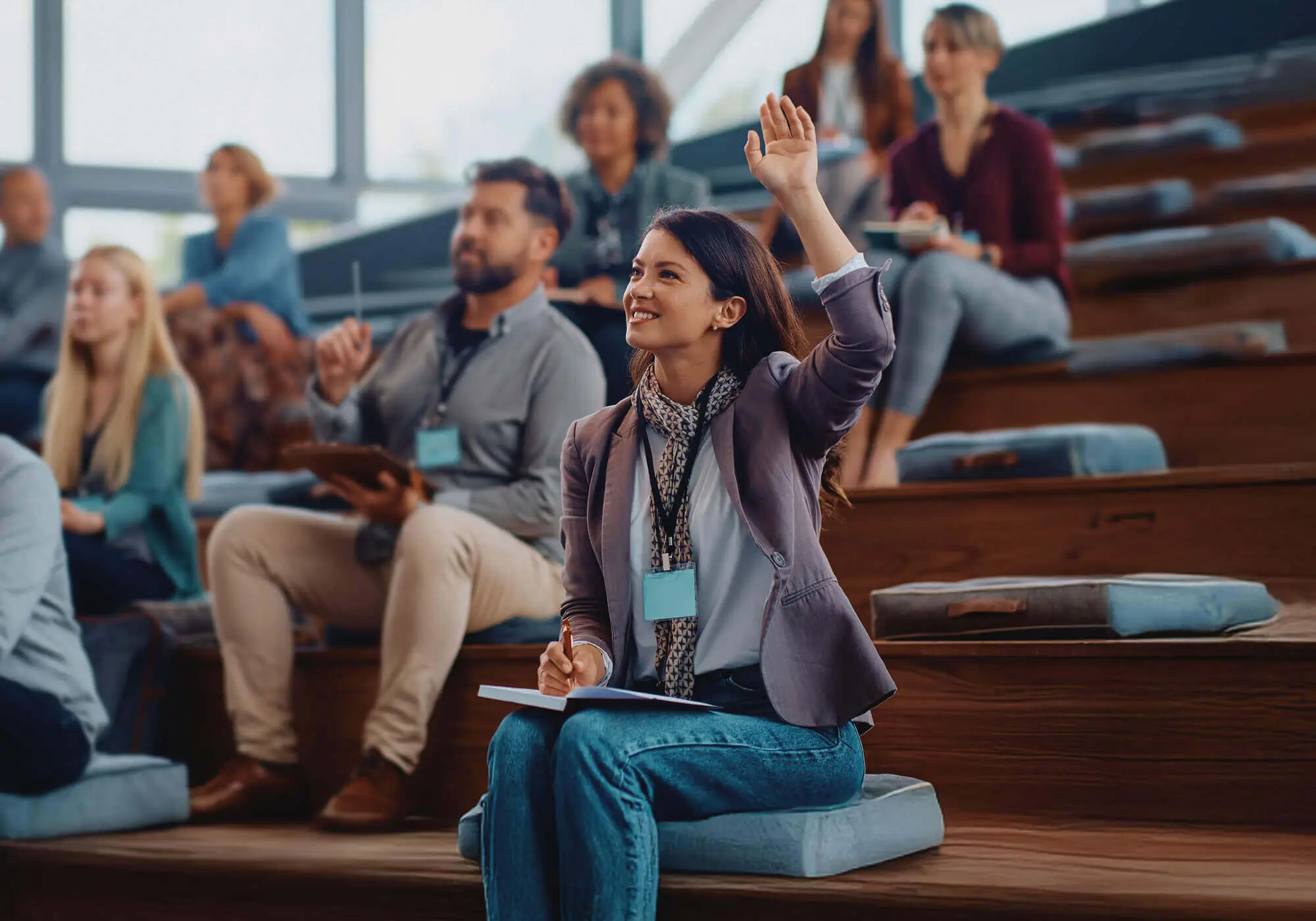 Mujer levantando la mano en una conferencia, participando activamente en un entorno de aprendizaje colaborativo. La sala cuenta con asistentes atentos, tomando notas y escuchando al ponente en un auditorio moderno con iluminación natural. Conceptos clave: educación, networking, formación profesional y desarrollo empresarial.