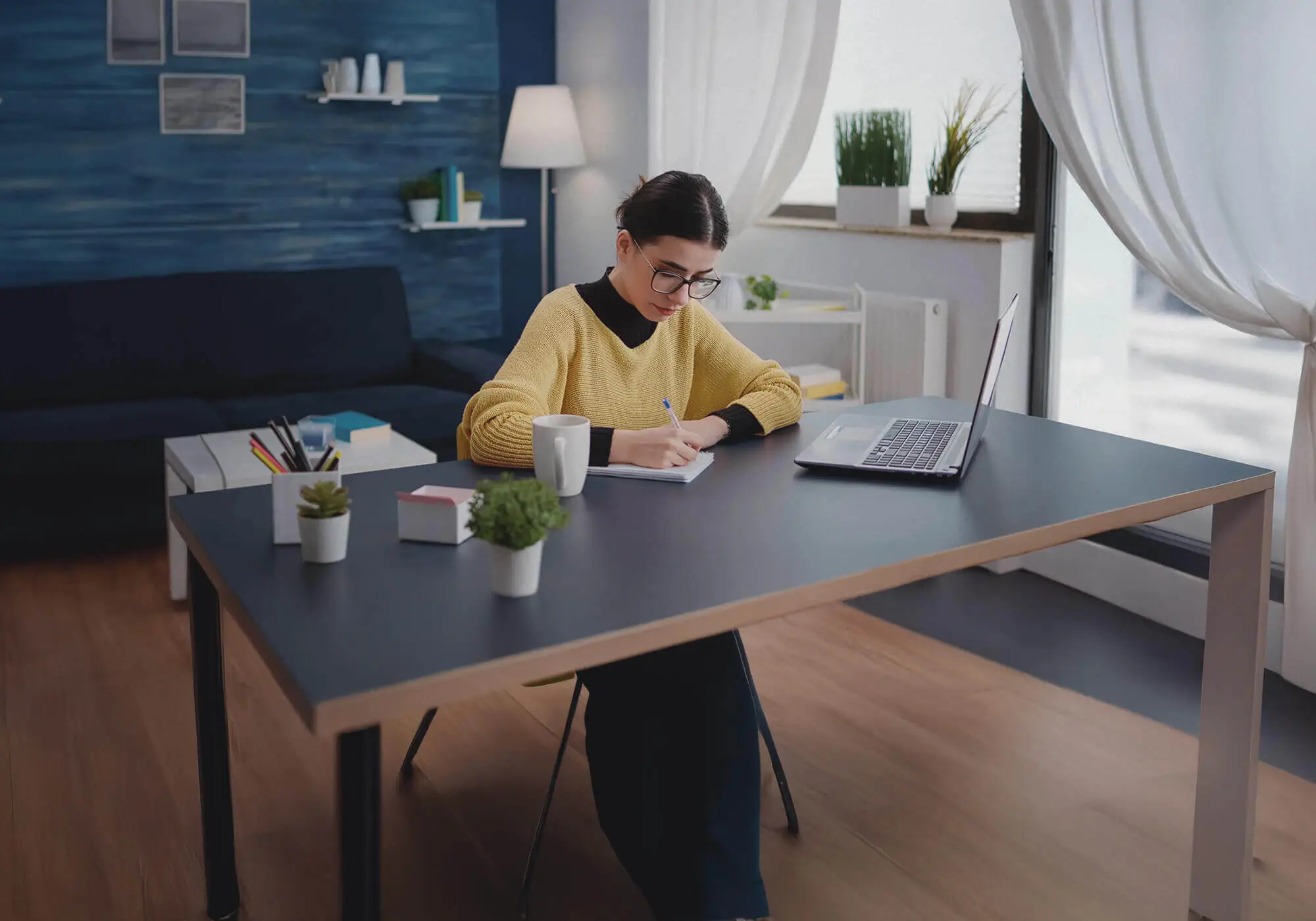 Mujer joven estudiando desde casa en un ambiente acogedor y moderno. Está sentada en un escritorio con una taza de café, cuaderno y portátil abierto, escribiendo con concentración. El fondo muestra una decoración elegante con estanterías, plantas y una pared azul, resaltando el concepto de teletrabajo, productividad y aprendizaje en un entorno inspirador