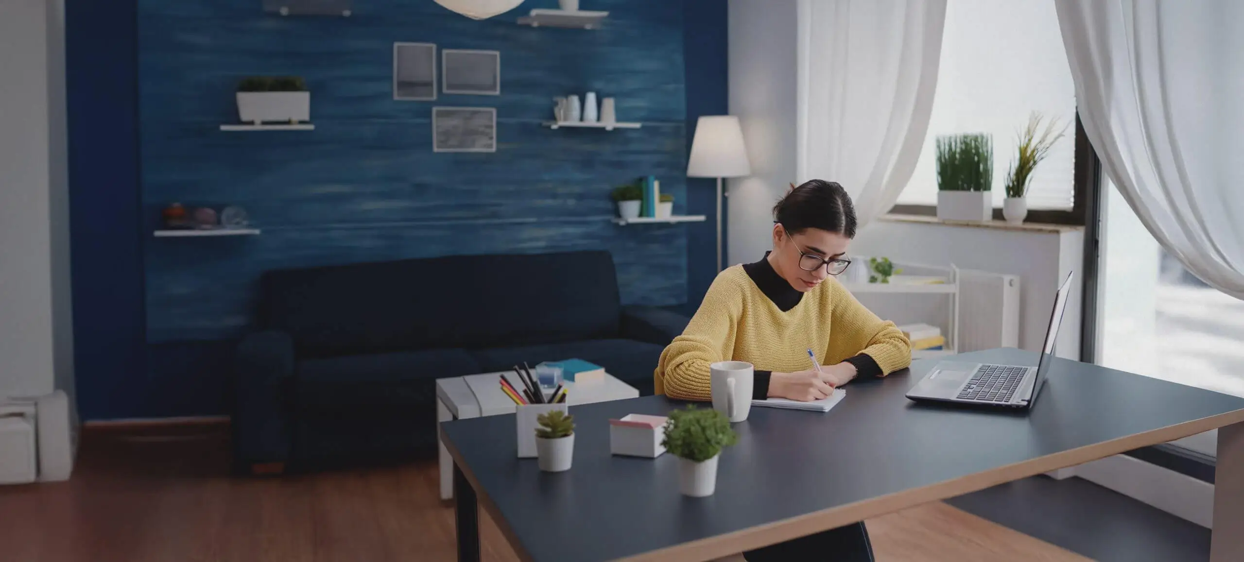 Mujer joven estudiando desde casa en un ambiente acogedor y moderno. Está sentada en un escritorio con una taza de café, cuaderno y portátil abierto, escribiendo con concentración. El fondo muestra una decoración elegante con estanterías, plantas y una pared azul, resaltando el concepto de teletrabajo, productividad y aprendizaje en un entorno inspirador